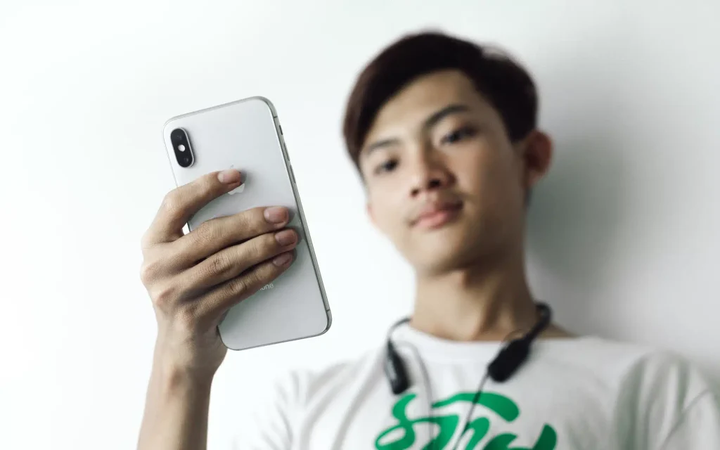 A young man smiling and holding up his iPhone while wearing headphones.